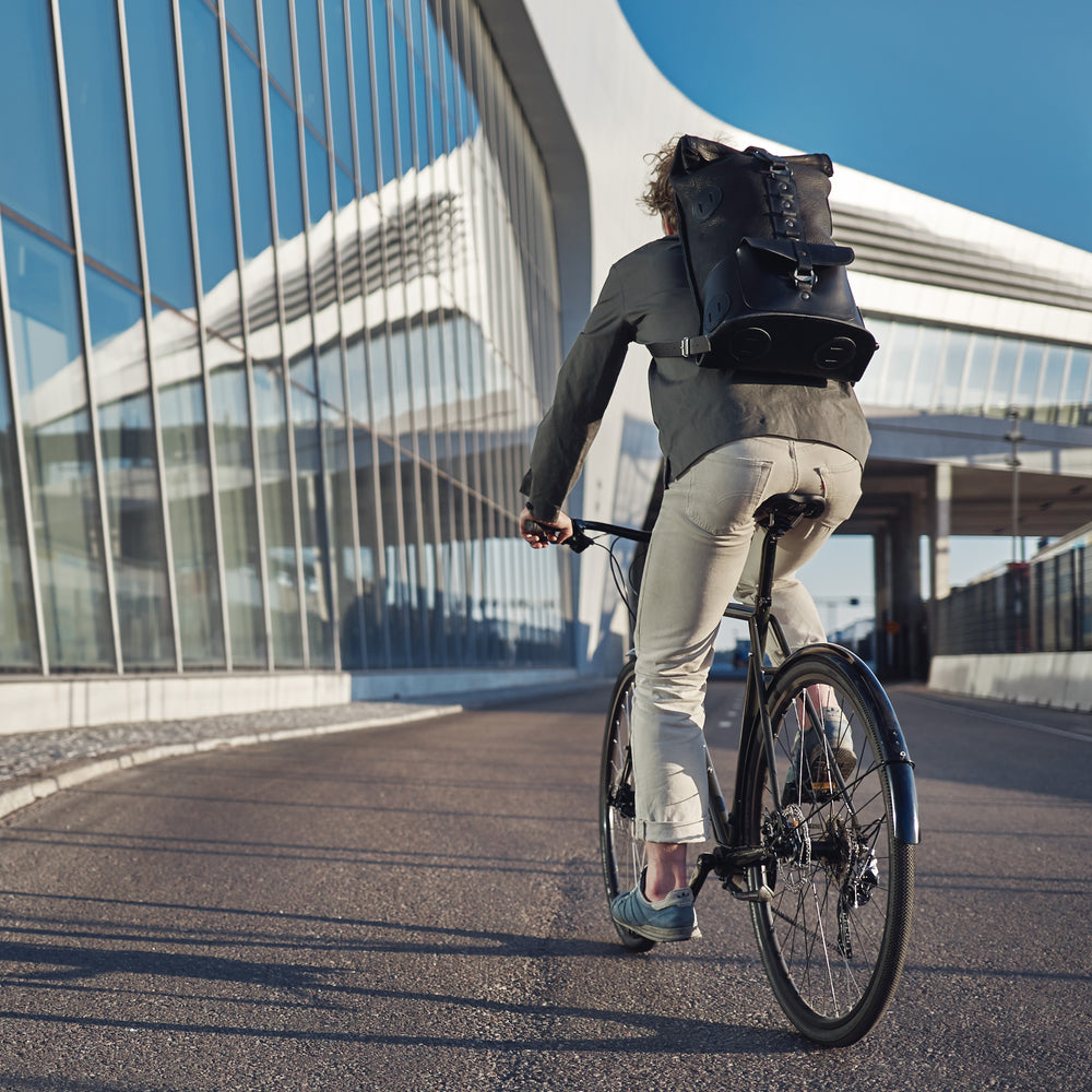 Black Fold Top leather backpack – worn by a cyclist riding past a modern glass building